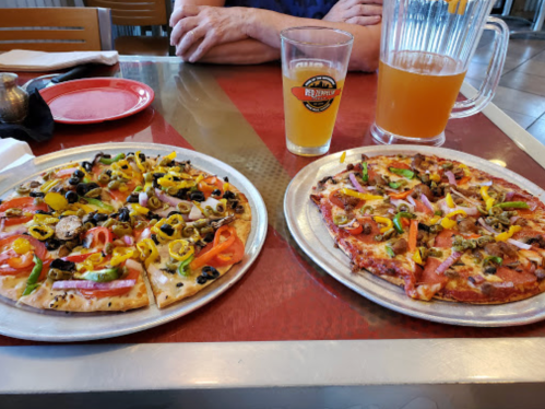 Two pizzas on metal plates: one topped with vegetables and olives, the other with meats and veggies, alongside drinks.