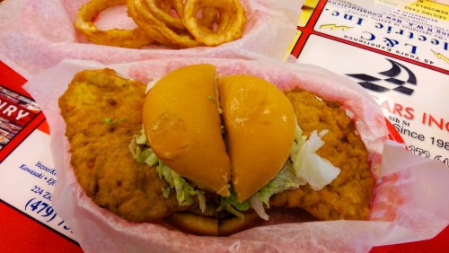A fried fish sandwich with lettuce on a bun, served with onion rings in the background.