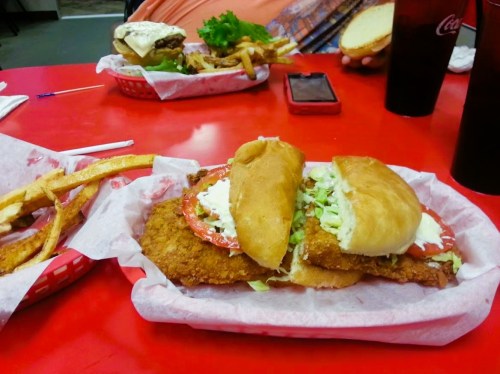 A close-up of a fried chicken sandwich with lettuce and tomato, served with fries and drinks on a red table.