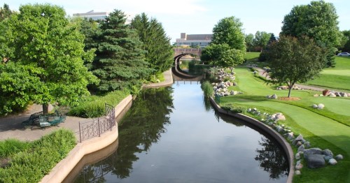 A serene landscape featuring a calm river, lush greenery, and a bridge in a park setting.