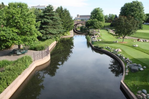A serene park scene featuring a winding canal, lush greenery, and trees reflecting in the calm water.