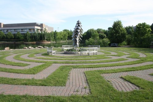 A circular labyrinth made of grass and bricks, featuring benches and a decorative sculpture in the center.