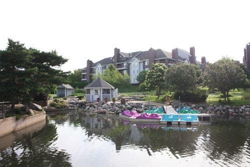A serene lake scene with colorful paddle boats, a gazebo, and lush greenery surrounding a residential building.