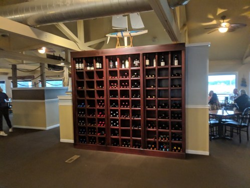 A wooden wine rack filled with various bottles in a restaurant setting, with diners visible in the background.
