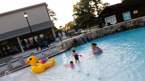A family enjoys a pool day with a large rubber duck, kids playing, and a woman in the water near a waterfall.