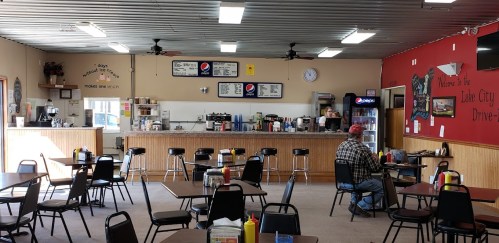 A cozy diner interior with tables, a counter, and a menu board, featuring a customer and a staff member.