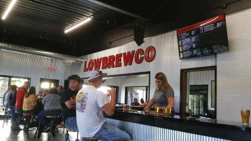 Interior of a brewery with patrons at the bar, a woman serving drinks, and a menu displayed on the wall.