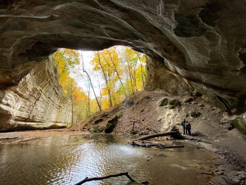 A person stands near a river inside a large cave, surrounded by autumn trees and rocky cliffs.