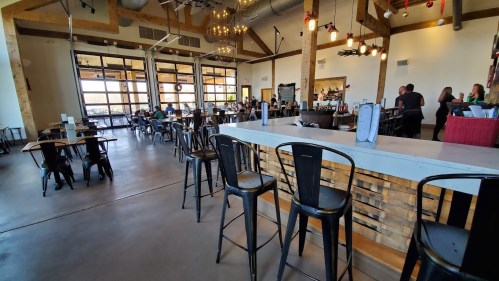 A spacious restaurant interior with wooden beams, a bar area, and patrons dining at tables. Bright and inviting atmosphere.