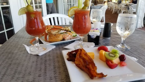 A table setting with two cocktails, a seafood dish, and a plate of fruit and bacon. Bright and inviting atmosphere.