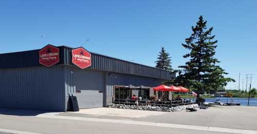 Exterior of Lake & Woods Brewing Company with outdoor seating, red umbrellas, and a view of the water nearby.