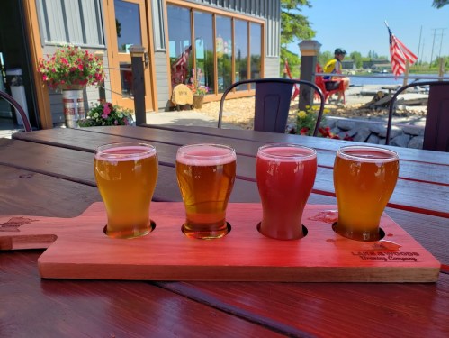 A wooden flight paddle holds four glasses of different craft beers on a table, with a sunny outdoor setting in the background.