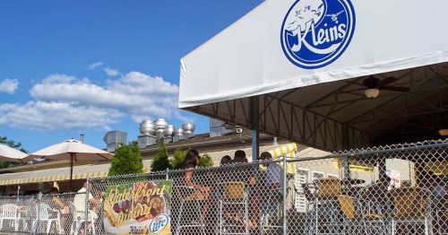 Outdoor dining area at Klein's, featuring a tent, tables, and a fence, with a blue sky and clouds in the background.