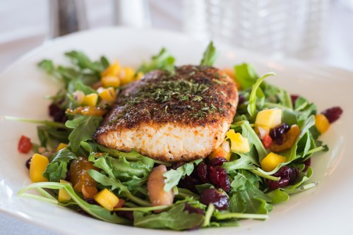 A plate of arugula salad topped with a seasoned salmon fillet and mixed fruits, garnished with herbs.