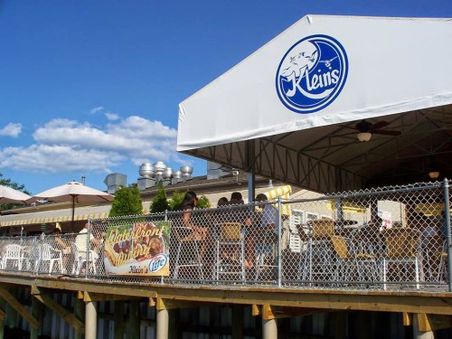 Outdoor dining area of a restaurant with a large tent, tables, and a sign reading "Klein's." Blue sky in the background.