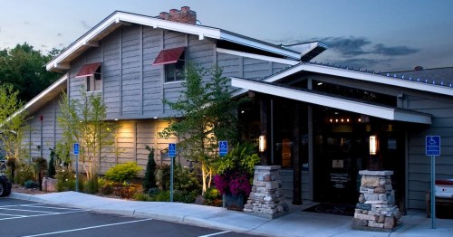 A modern, rustic building with a sloped roof, surrounded by greenery and parking spaces, illuminated at dusk.