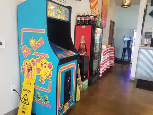 A colorful Mr. Pac-Man arcade machine next to a soda fridge in a casual dining area. Caution sign on the floor.