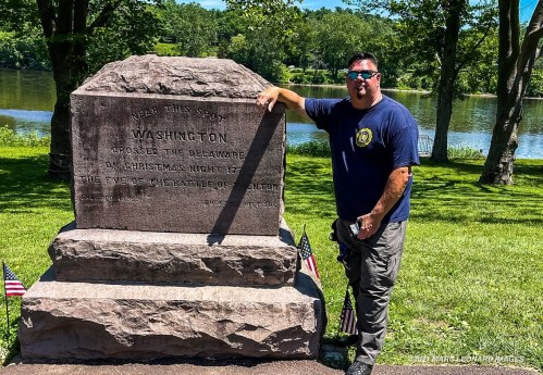 A man stands beside a historical monument commemorating Washington's crossing of the Delaware River, with flags nearby.