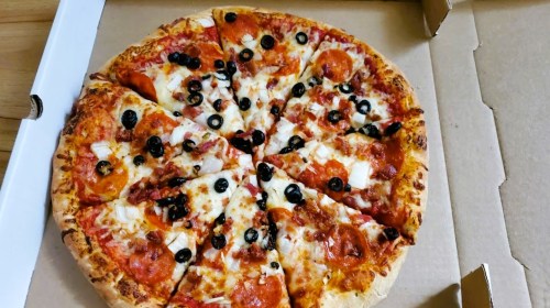 A pizza with pepperoni, black olives, and cheese, sliced into wedges, in a cardboard pizza box.