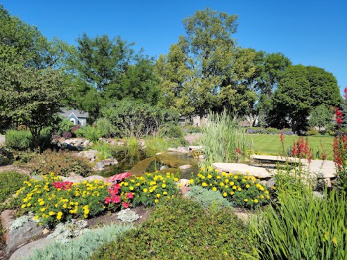 A vibrant garden with colorful flowers, lush greenery, and a serene pond under a clear blue sky.