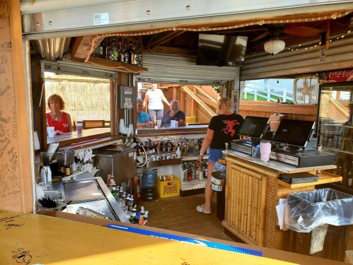 A beach bar with a bartender serving drinks, surrounded by shelves of liquor and patrons enjoying the atmosphere.