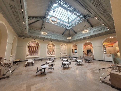 A spacious, well-lit hall with high ceilings, large windows, and tables arranged for seating. Colorful artwork on the walls.