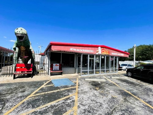 A barbecue restaurant with a large dinosaur statue outside, featuring a red awning and a parking lot.