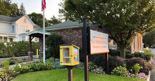A cozy coffee shop with a colorful little free library and a flag, surrounded by flowers and greenery.