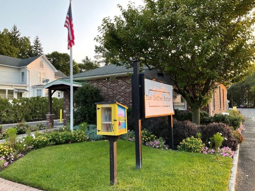 A cozy coffee shop with a colorful little free library, surrounded by flowers and an American flag in the background.