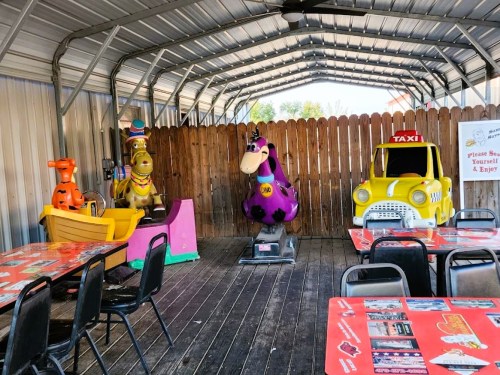 Colorful kiddie ride machines in a covered outdoor area with tables and chairs.