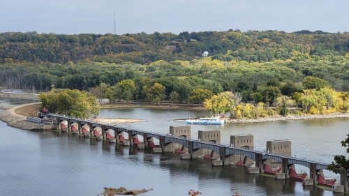 A scenic view of a river with a dam, surrounded by trees in autumn colors and a boat in the distance.