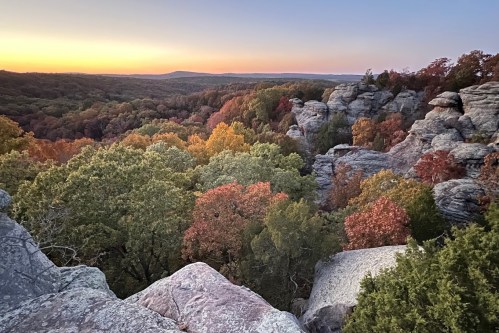 A scenic view of colorful autumn trees and rocky cliffs at sunset, with rolling hills in the background.