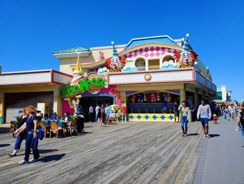 Colorful amusement arcade with clown decorations, people walking on a boardwalk, and clear blue skies.