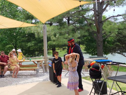 A magician performs for children outdoors, with an audience seated nearby under shaded canopies by a lake.