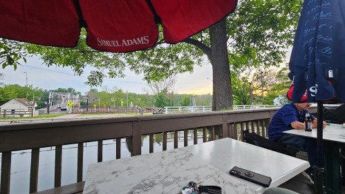 A view from a patio with a Samuel Adams umbrella, overlooking a river and trees at sunset. A person is seated nearby.