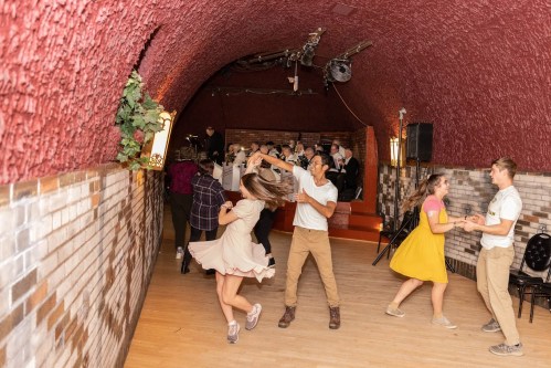 A lively dance scene in a rustic venue, with couples twirling and a band playing in the background.