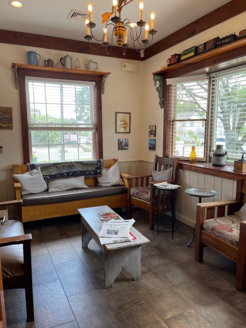 Cozy café interior with wooden furniture, a chandelier, and natural light from windows. Relaxed seating area.