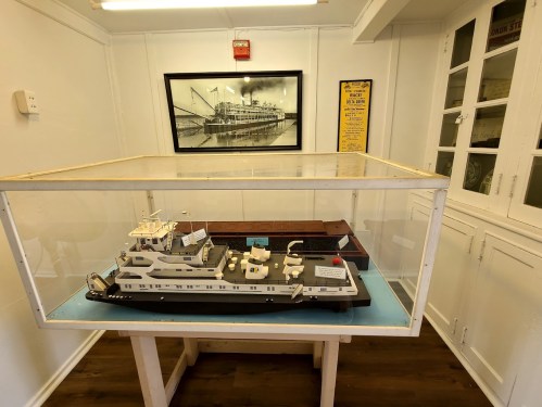 A model of a boat displayed in a glass case, with a black-and-white photo of a steamboat on the wall behind it.