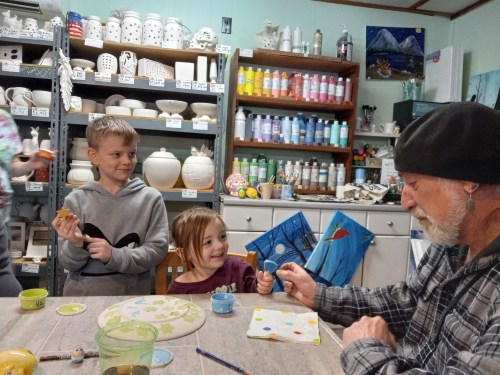 Two children and an older man are painting pottery in a colorful art studio filled with supplies and finished pieces.