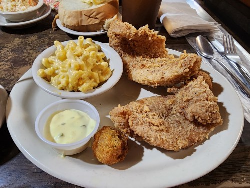 A plate of fried chicken, macaroni and cheese, a cornbread muffin, and dipping sauce, with bread in the background.