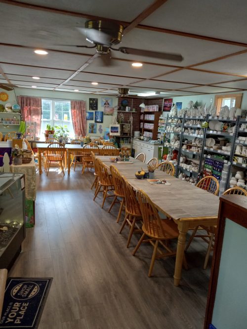 A spacious, well-lit art studio with wooden tables, chairs, and shelves filled with pottery and art supplies.
