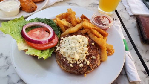 A plate with a burger topped with blue cheese, lettuce, tomato, onion, and crinkle-cut fries, alongside dipping sauce.