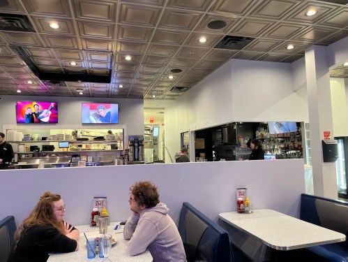 A diner interior with two women at a table, a counter, and TVs showing entertainment in the background.