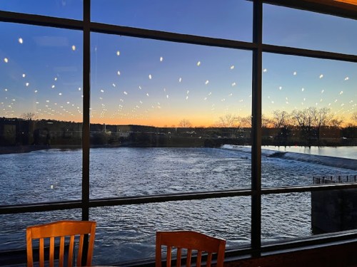 A serene sunset over a river, viewed through large windows with string lights reflecting in the glass.