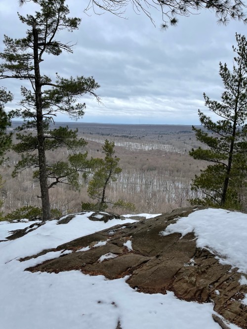 A rocky outcrop with patches of snow overlooks a vast, wooded valley under a cloudy sky.