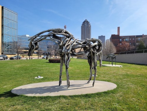 A wooden horse sculpture stands on a grassy area with a city skyline in the background under a clear blue sky.