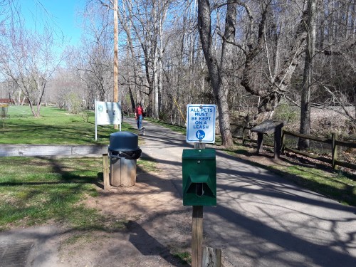 A park path with a trash can and a pet waste station, featuring a sign that says "All pets must be on a leash."