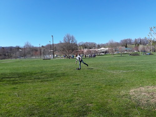 A person kicks a soccer ball on a grassy field with trees and a blue sky in the background.