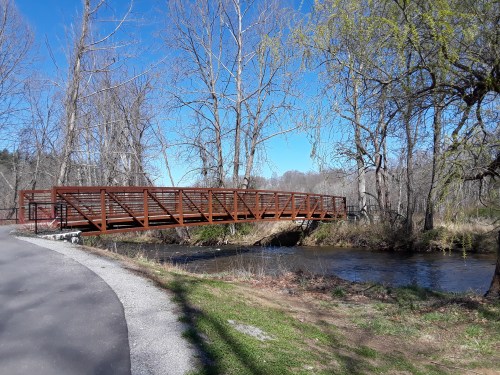 A metal footbridge spans a calm stream, surrounded by trees and a clear blue sky. A path runs alongside the water.