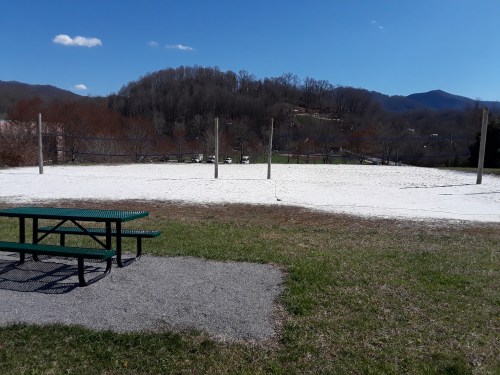 A sandy volleyball court with poles, surrounded by grass and mountains under a clear blue sky. A picnic table is nearby.